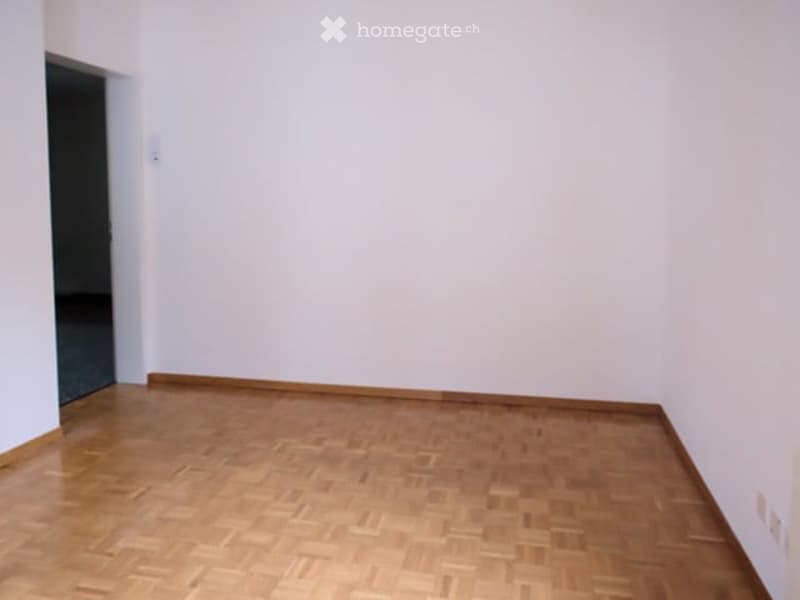 An empty room with white walls, wooden floor, and an open door leading to another room.