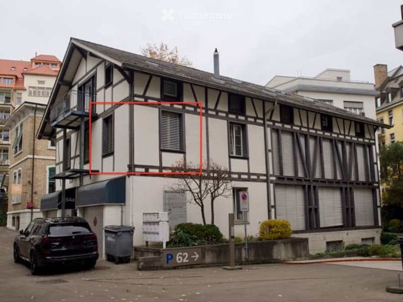 A three-story building with a red square highlighting a specific apartment on the second floor. The building has a mix of white and brown exterior walls, with a balcony on the left side. A black car is parked in front of the building, and a trash bin is p