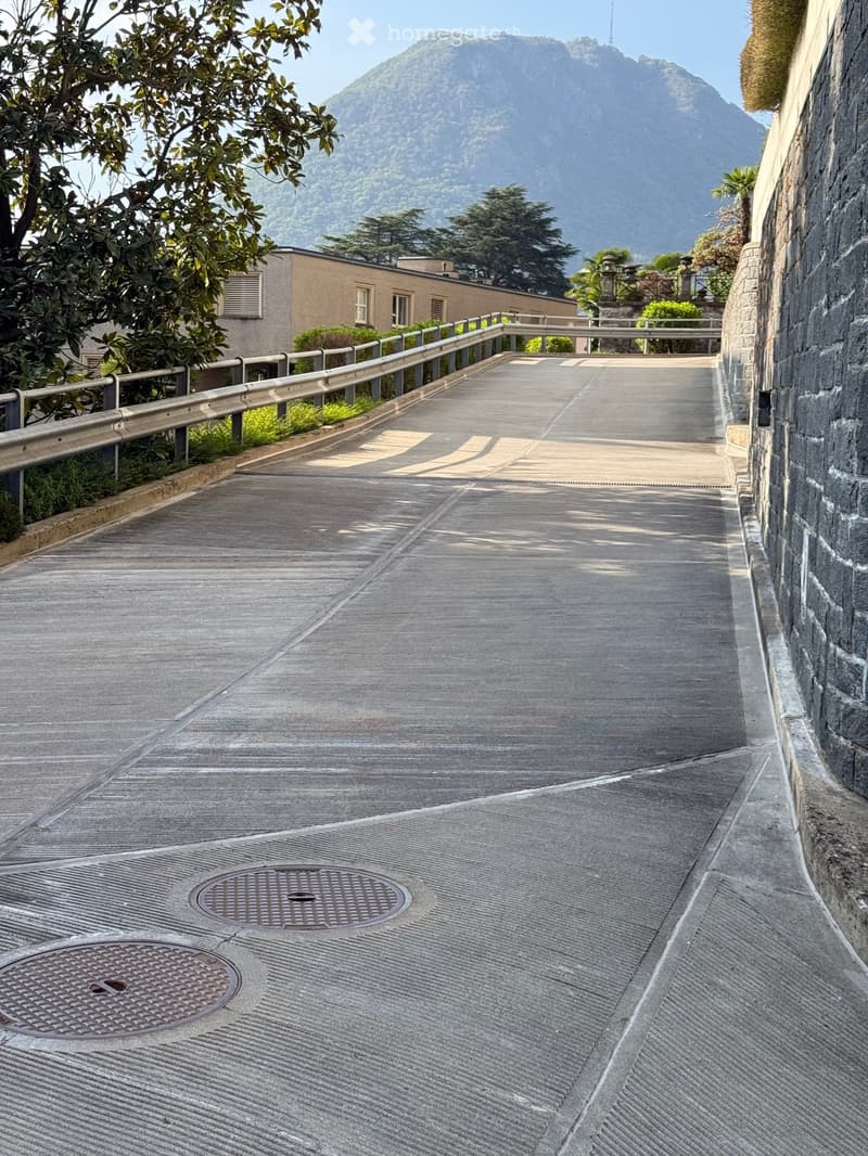 Corridor / Hallway. A concrete pathway with a metal railing on the left side, two manhole covers, and a stone wall on the right. The pathway is empty, and there is a building with a tower in the distance.