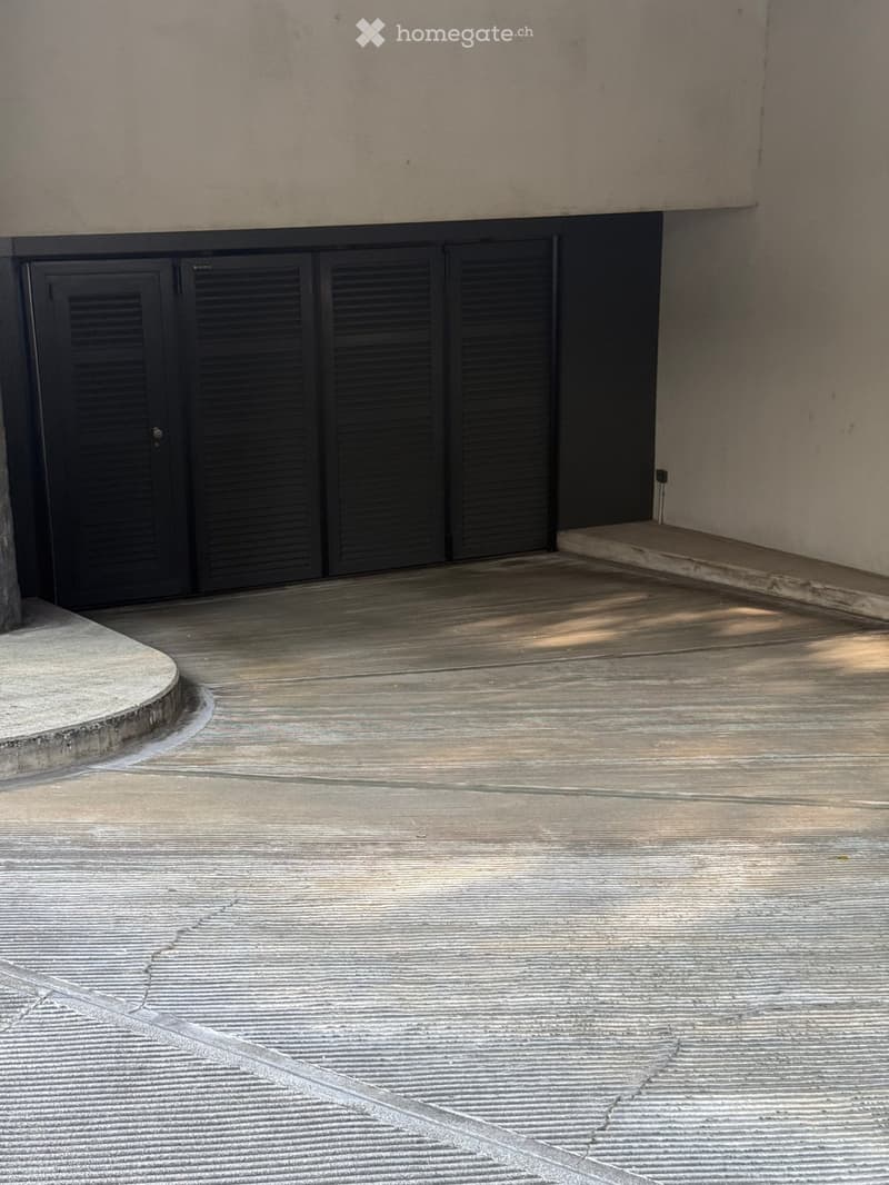 Unfurnished Room. An empty room with a concrete floor and a wall with black shutters. The floor is clean and has a crack in the middle. The wall is white and has a power outlet on the right side. The room is probably a garage or a storage room.