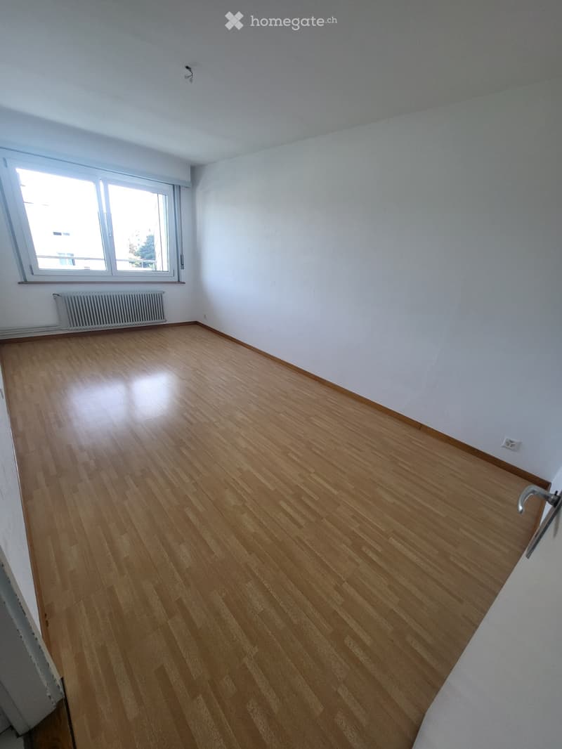 Empty room with wooden flooring, white walls, and a window with blinds. A radiator is visible below the window, and the room has a clean, minimalist design.