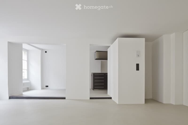 An empty room with white walls, ceiling, and floor. There is a window on the left side and a small cabinet on the right side. The room has a modern design with a sleek and minimalist look.
