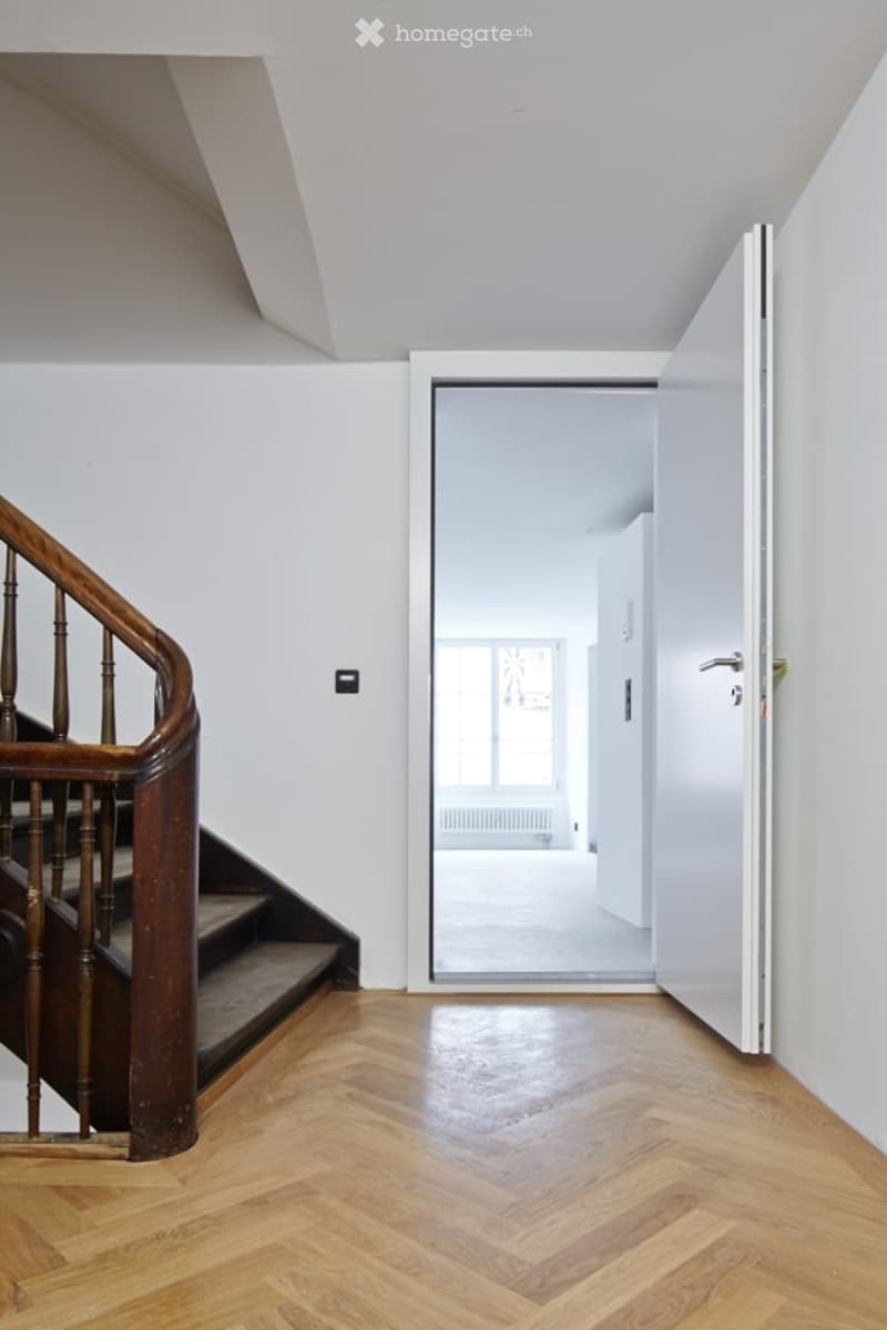 A corridor with a wooden staircase leading to the upper floor, a white door, and a white wall.