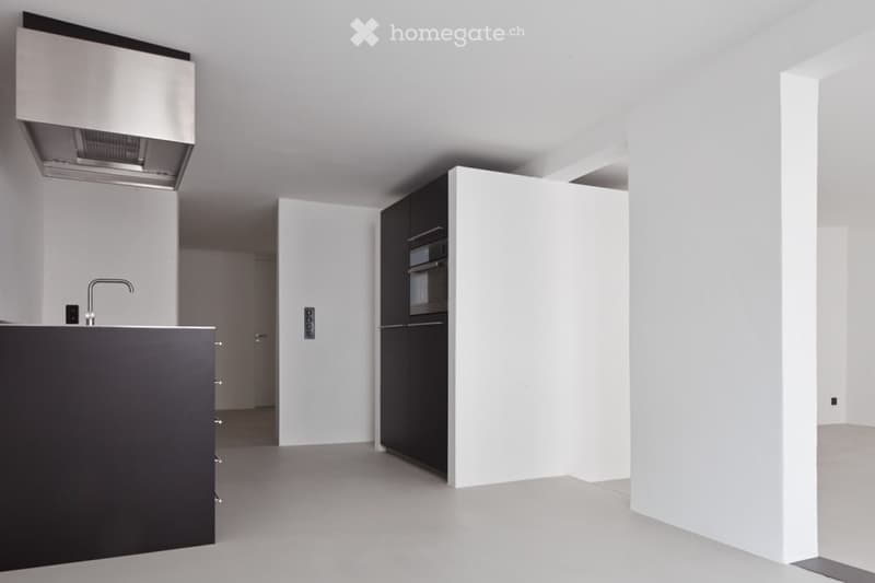 An empty kitchen with white walls, gray flooring, a black kitchen island, and a stainless steel range hood. There is a faucet and a power outlet on the wall.