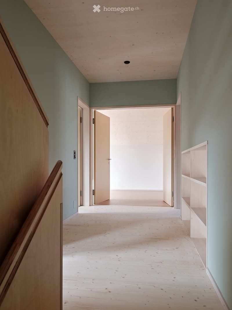 Empty corridor with light blue walls and wooden floor. Two open doors lead to another room. A wooden handrail is on the left side.