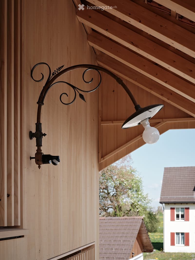 A vintage-style outdoor light fixture mounted on a wooden wall, featuring a curved metal arm and a white light bulb.