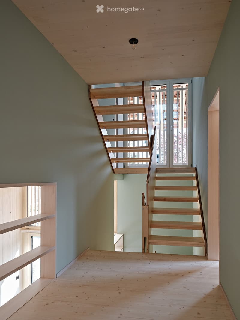 An empty room with wooden floors and walls painted in a light green color. A wooden staircase is positioned in the center of the room, leading to an upper level. The staircase has a modern design with a wooden handrail and steps. There are two glass doors