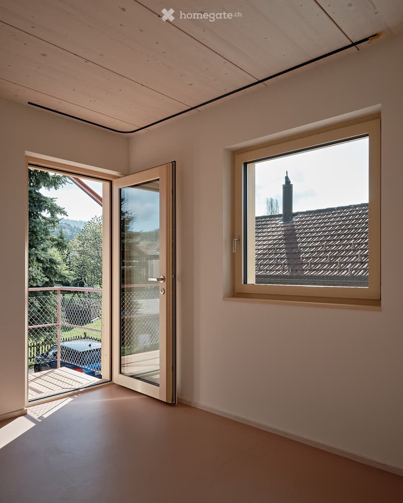 Empty room with wooden ceiling, white walls, and large windows. One window offers a view of a house with a chimney and trees.