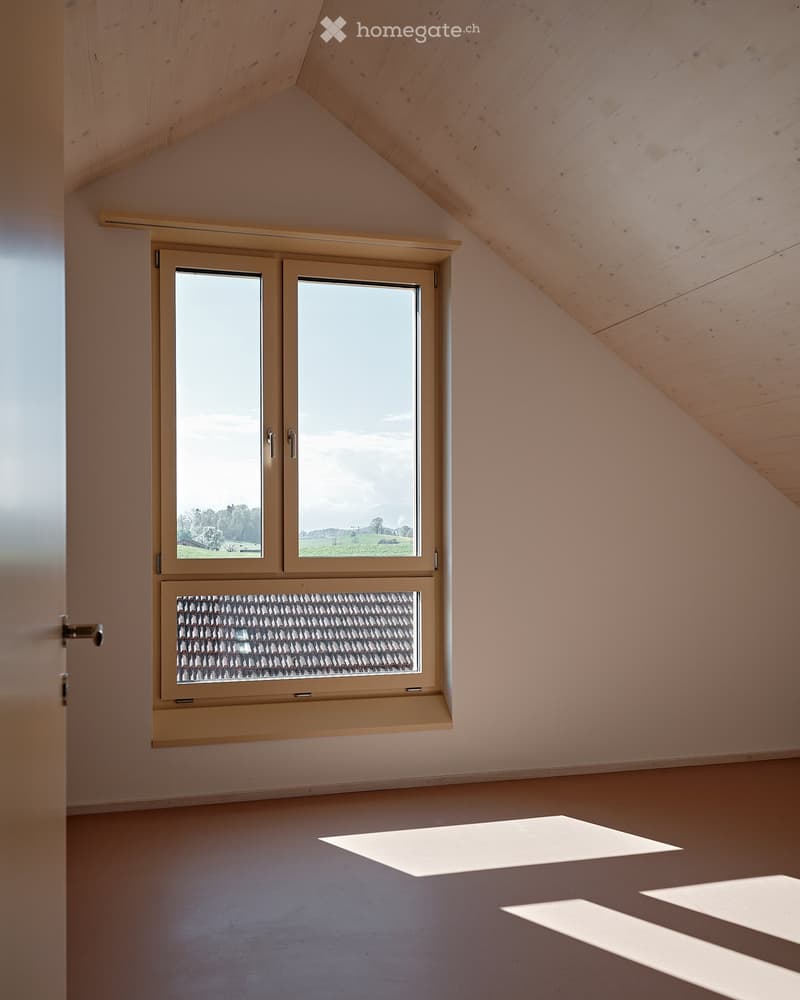 An empty attic room with a window and wooden ceiling. The room has a white wall and a brown floor.