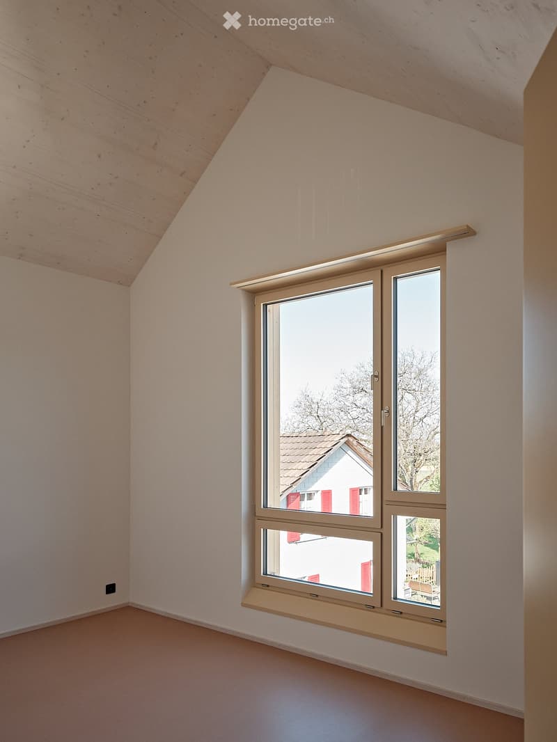 Empty room with white walls, wooden ceiling, and a large window overlooking a house with red shutters.