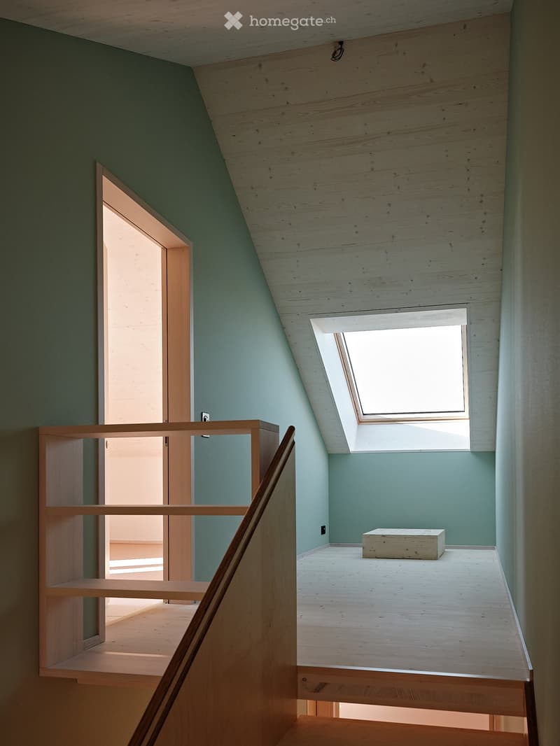 An attic room with wooden ceiling, green walls, and a wooden box on the floor. The room has a wooden handrail and a wooden shelf on the left side. There is a door on the left side and a window on the right side. The room is empty and has no furniture.