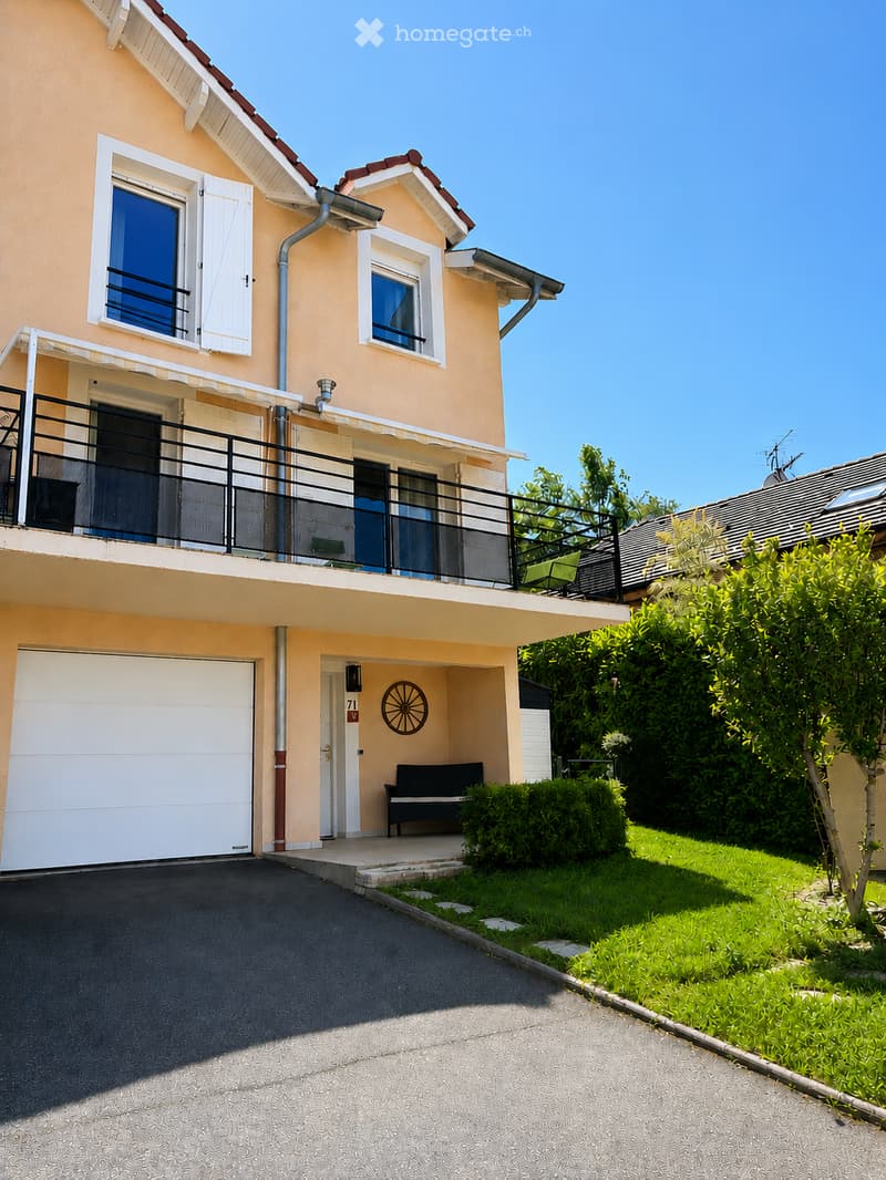 Two-story house with beige exterior, white-framed windows, garage, balcony, and small garden area.
