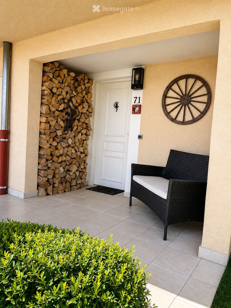 A cozy outdoor seating area with a wicker chair and cushion, a neatly stacked woodpile, a decorative wheel wall art, and a white front door with the number 71.