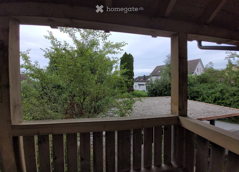 Balcony with wooden railing, overlooking a green, tree-filled area with houses in the background.