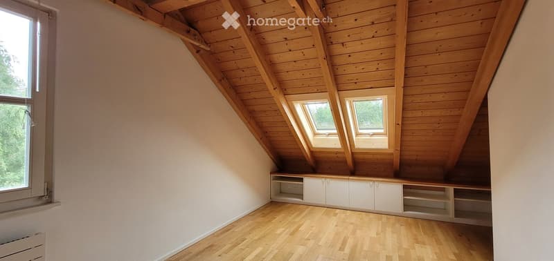 empty attic room with wooden ceiling, wooden floor, white walls, two windows, built-in cabinets, radiator