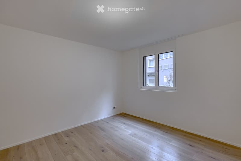 Completely empty room with white walls and wooden floor. A window is on the right wall, and there is a power outlet on the wall near the window.