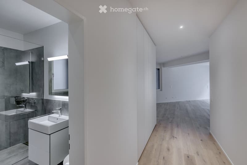 Modern bathroom with white walls, wooden floor, and a glass shower enclosure. Features a white sink with a faucet, a mirror, and a white toilet. The shower area is equipped with a shower head and a glass door. The room is well-lit with recessed ceiling li