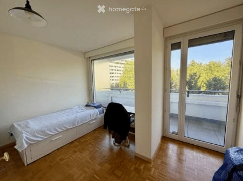 A small furnished room with a bed, chair, desk, and lamp. The room has wooden floors and large glass doors leading to a balcony. There is a blue bag on the floor.