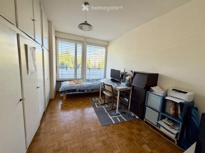 A small furnished room with a bed, desk, and various office equipment. The room has wooden floors and white walls.