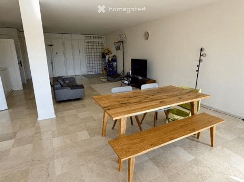 Spacious room with wooden dining table, bench, and chairs. A couch is placed near the wall with a television and a lamp. A basketball hoop is mounted on the wall, and a rug is on the floor. A clock is also mounted on the wall.