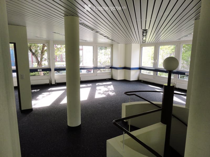 Large open space, white walls, black carpeting, glass windows with blue trim, central column, staircase at the corner