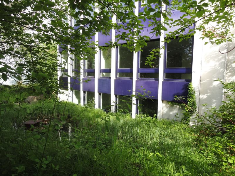 A modern office building with multiple floors, characterized by its white and blue facades, featuring many windows, and surrounded by greenery.