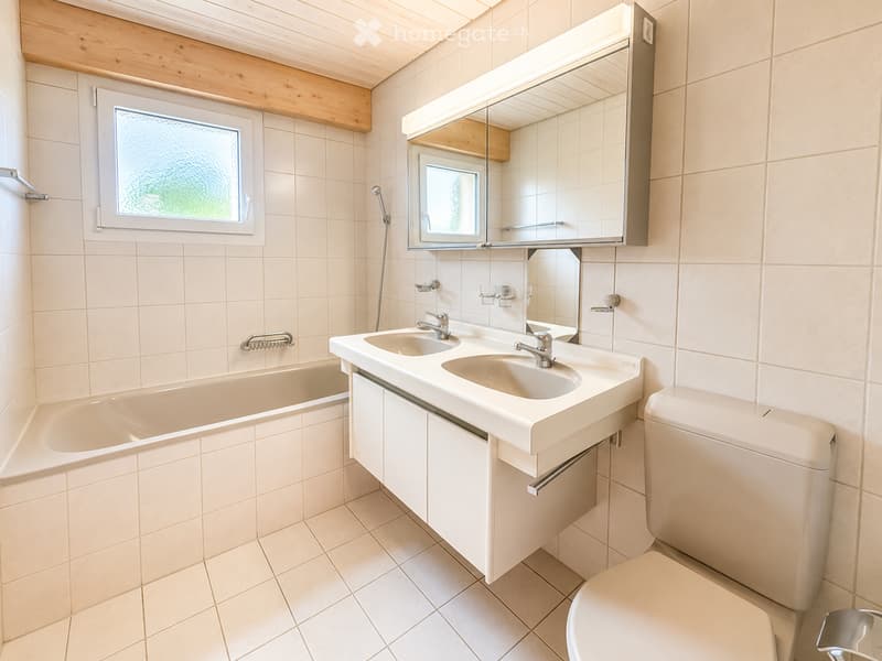 Modern bathroom with a bathtub, double sink, toilet, and a large mirror with built-in lighting. The room features light-colored tiles and a window with frosted glass.