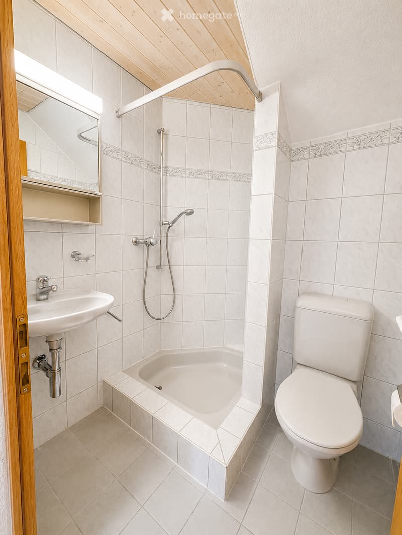 Modern bathroom with white tiles, a bathtub, toilet, sink, and a showerhead. The room features a wooden door frame and a built-in shelf above the sink.