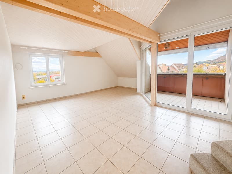 Bright, unfurnished attic room with wooden slanted ceilings, large windows, and a sliding glass door leading to a balcony with scenic views.