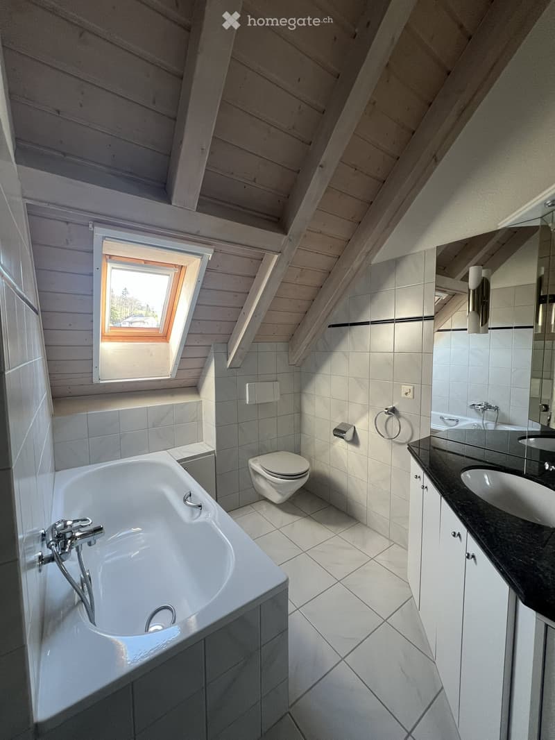Bathroom with a bathtub, toilet, sink, and mirror. The walls and floor are tiled in white. There is a wooden ceiling with a skylight window.