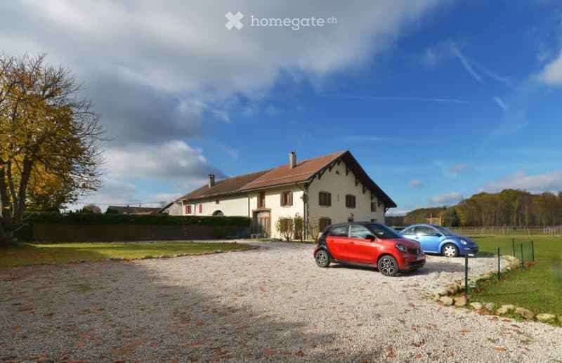 House. A two-story house with a brown roof and white walls, surrounded by a gravel driveway and a grassy area. Two cars are parked in front of the house. The house is located in a rural area with trees and a fence in the background.