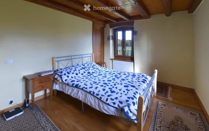 Bedroom. A bedroom with a bed, nightstand, and rug. The bed has a blue and white patterned bedspread. The nightstand has a book and a cell phone on it. The room has wooden flooring and white walls. There is a window with a view of a field and trees outsid