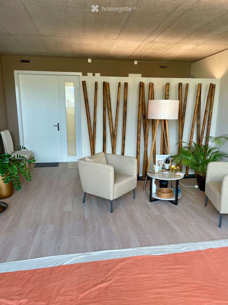 A well-lit room with a bed, two chairs, a small round table, a floor lamp, and several potted plants. Bamboo stalks are arranged against the wall, and a white door is visible in the background.