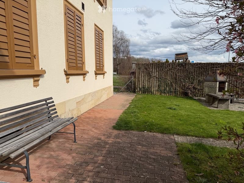A brick walkway leads to a wooden bench, with a house featuring three wooden shutters and a stone wall. Behind the house, there is a wooden fence with a fireplace and a bench.