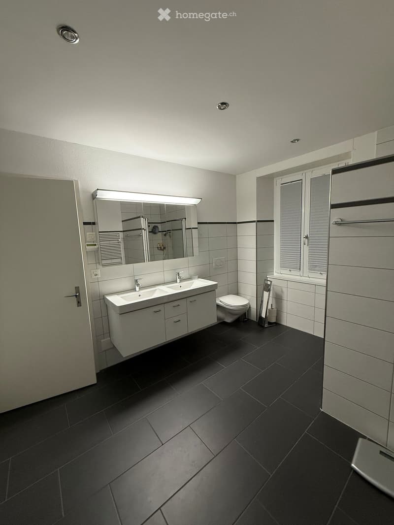 modern bathroom with double sink, toilet, large mirror, black tiles, white walls, window with blinds