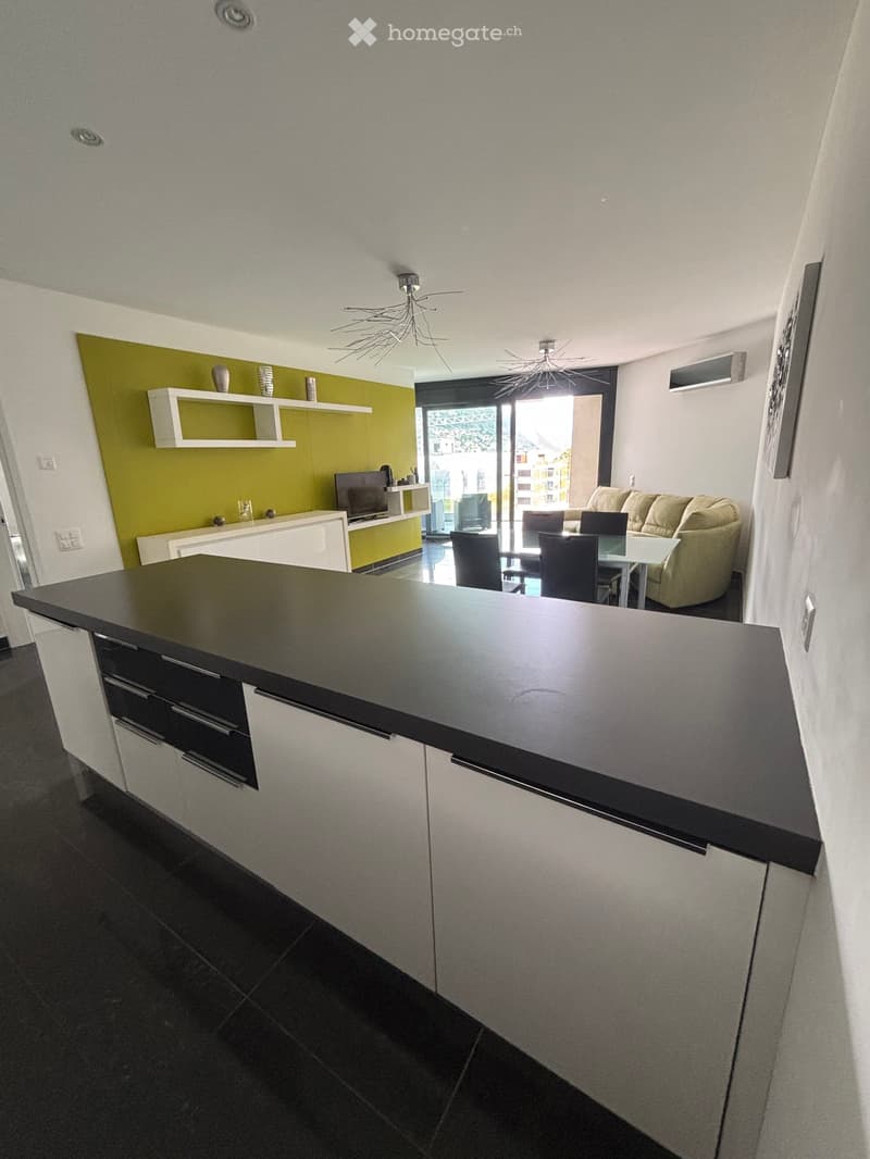 Modern open-plan living and dining area with a kitchen island, white cabinets, dark countertops, and a sleek design. Features a beige sofa, dining table with chairs, and a TV on a white stand.