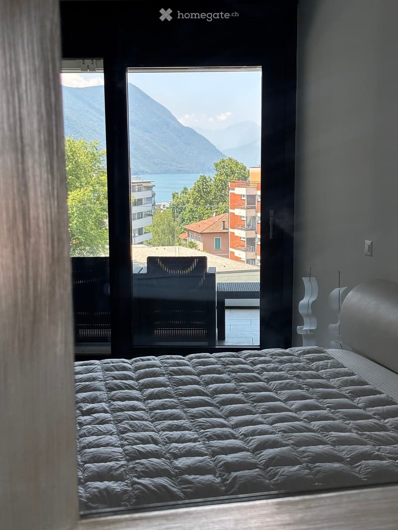 Bedroom with a bed, a chair, a lamp, and a bench. The room has a glass door leading to a balcony with a view of the mountains and the sea.