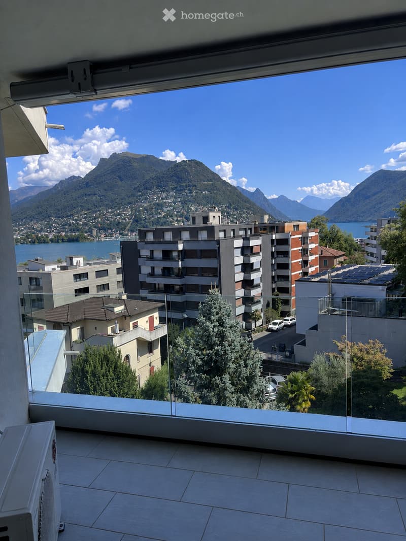 Balcony with a panoramic view of a lake and surrounding mountains, featuring modern glass railings and tiled flooring.