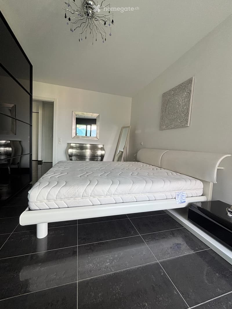 Modern bedroom with a white bed, mirrored dresser, wall art, and a decorative chandelier.