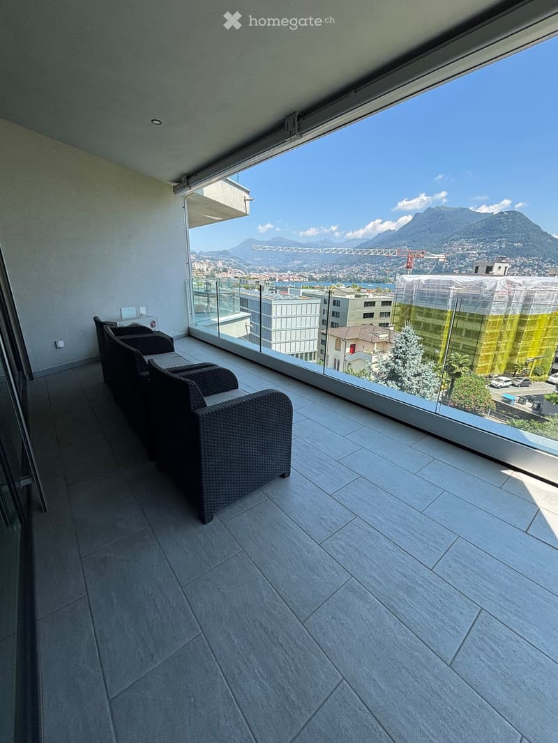 A balcony with a view of the city and mountains, featuring a tiled floor and a set of black chairs and a table.