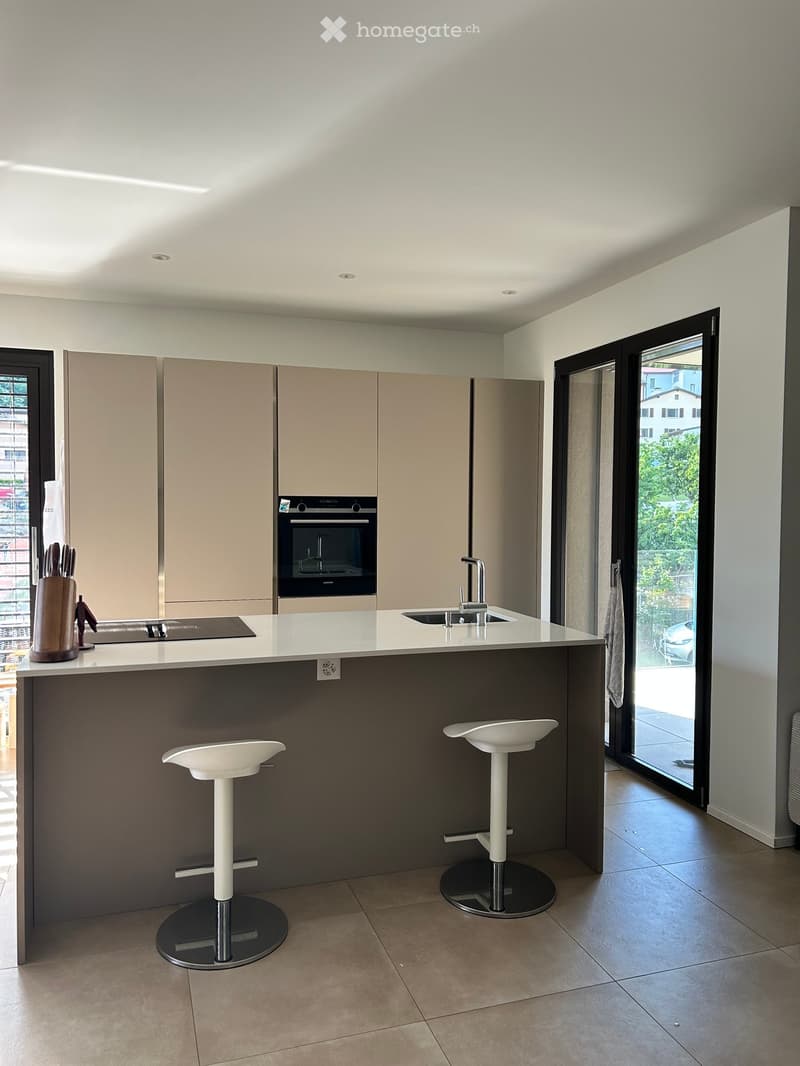 Modern kitchen with a kitchen island, stools, sink, faucet, oven, cabinets, cutting board, knives, power outlet, and dish towel.