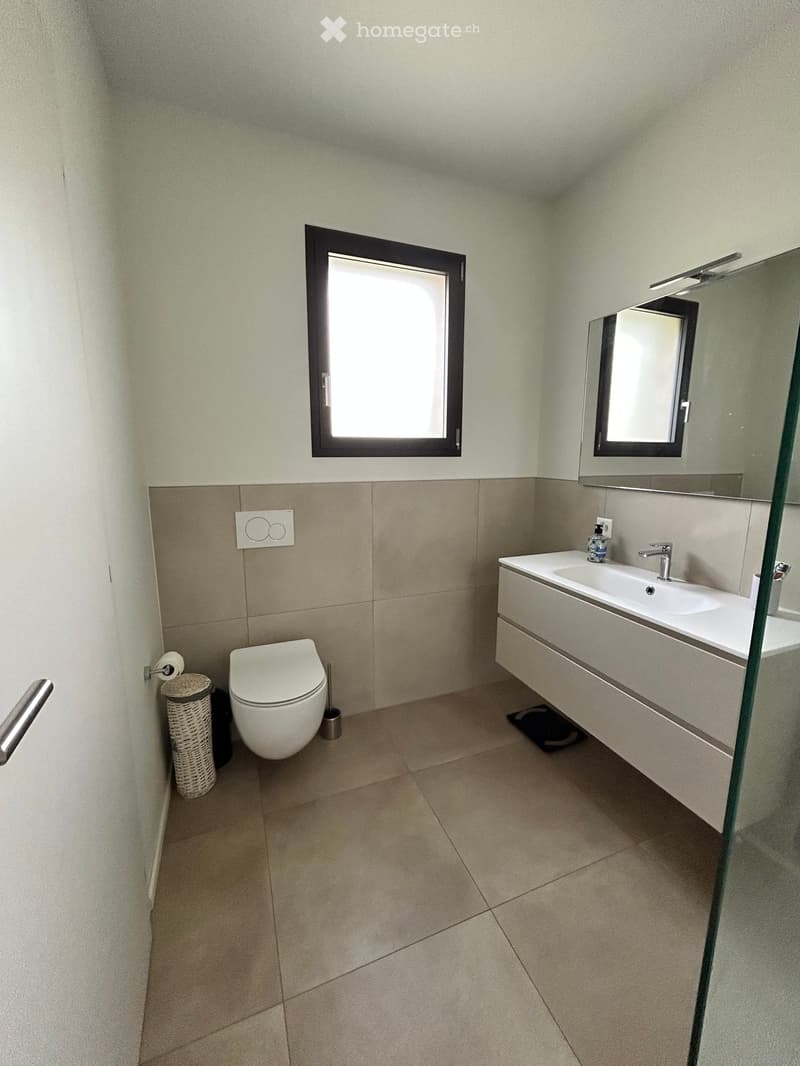 Modern bathroom with white tiles, a toilet, a sink, and a mirror. The sink has a faucet and a soap dispenser. There is a toilet brush and a basket with toilet paper next to the toilet.