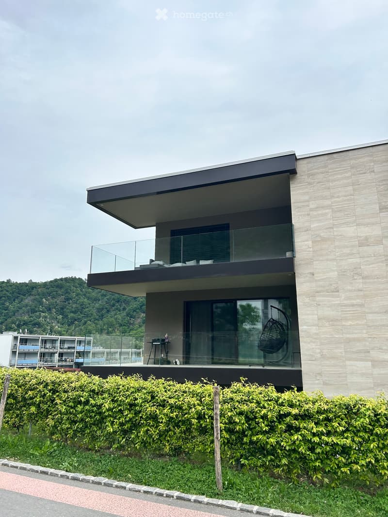 Modern building with glass balconies, beige stone walls, and a green hedge in front.