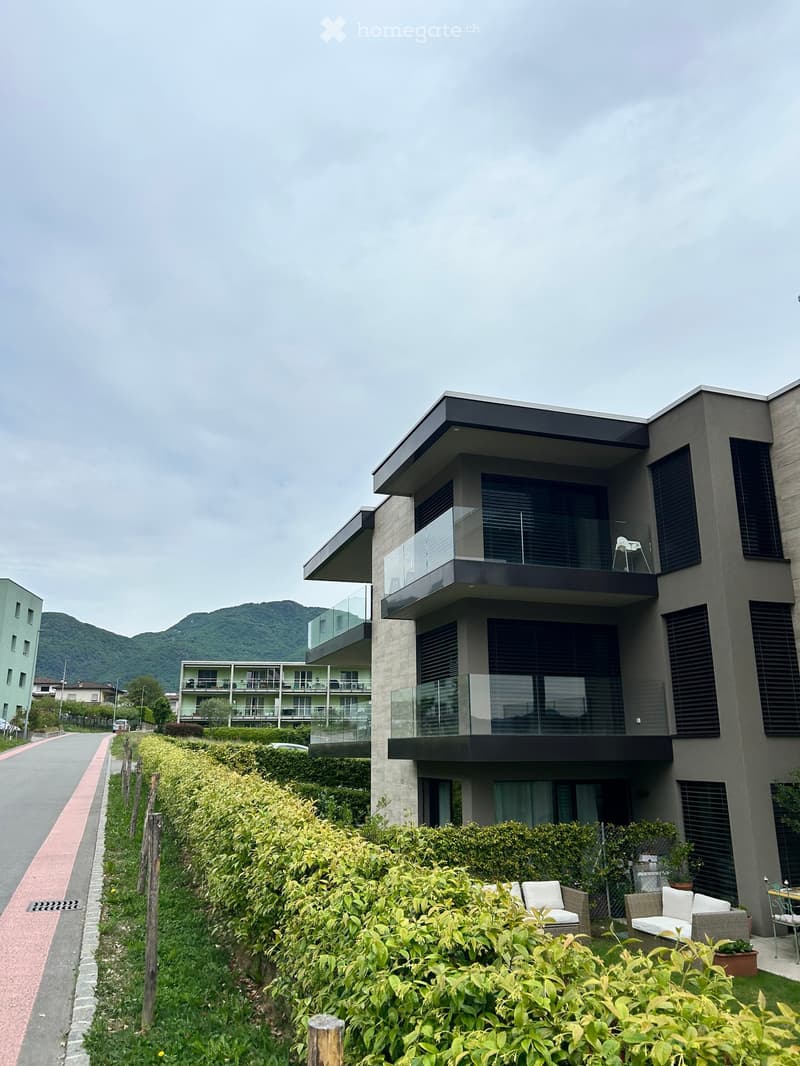 Modern building with glass balconies, surrounded by greenery and mountains in the background.
