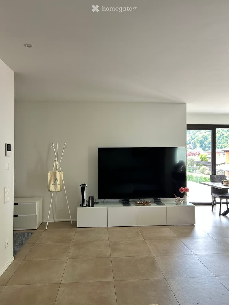 Modern living room with a large flat-screen TV on a white cabinet, a white coat rack, and a beige rug. The room has white walls and a tiled floor.