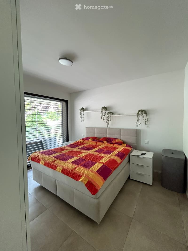 A bedroom with a bed, nightstand, and plants on the wall. The bed has a colorful bedspread and pillows. The nightstand has a drawer and a remote control. The window has blinds and a view of the outside. The ceiling has a light. The floor is tiled.