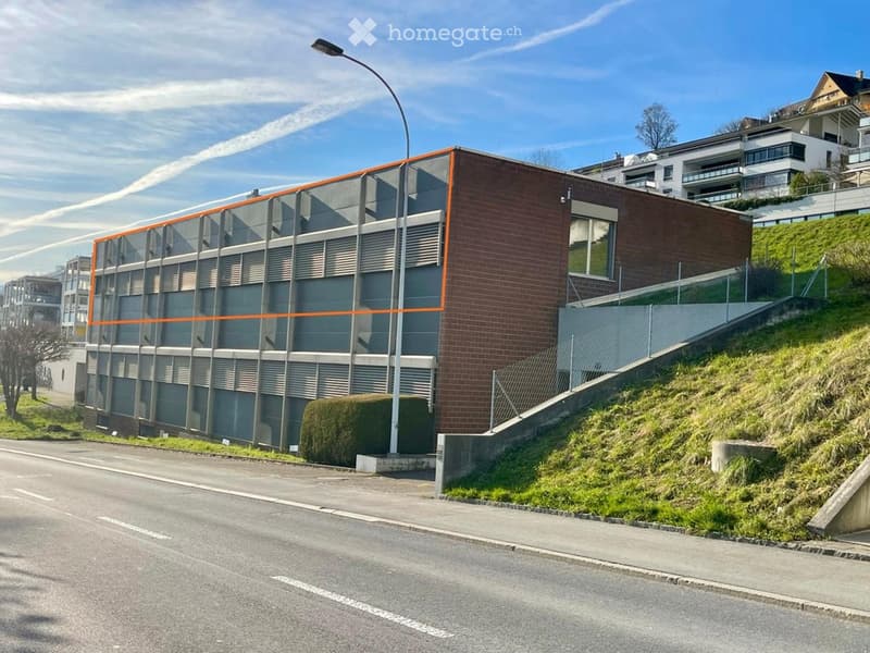 A multi-story building with a brick facade and glass windows, situated on a sloped area with a road in front.