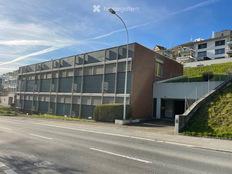 Modern apartment building with a gray facade, brick accents, and a glass wall. There is a driveway leading to the garage entrance with a slope and steps. The building is on a hillside with a lawn and streetlight. Other buildings are visible in the backgro