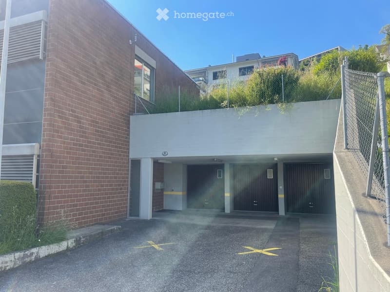 A building with a brick wall and a green roof, three garages, and a parking lot.