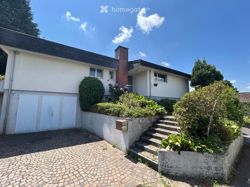 Two-story house with a flat roof, white exterior, and a brick chimney. Features a garage with a white door, a small garden with shrubs and flowers, and a paved driveway.